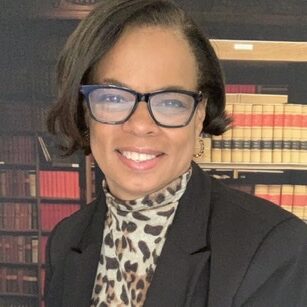 A woman in glasses and a jacket standing next to books.